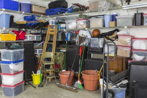 An interior storage area filled with various household rubbish and clutter, including clear plastic storage boxes with red, black, and blue lids, stacked on metal shelving units along the back wall. Several large empty terracotta flower pots are positioned in the foreground, with cleaning tools such as brooms and mops leaning against a small wooden step ladder and a metal trolley. A small wooden chair, a yellow bucket, and miscellaneous items such as bags and containers are scattered throughout the space. The environment appears to be a garage or storage room, well-lit with no natural light visible, and the floor is made of concrete. The scene depicts a typical cluttered space awaiting clearance, consistent with services offered by Harrow House Clearance for rubbish removal and waste disposal in the local area.