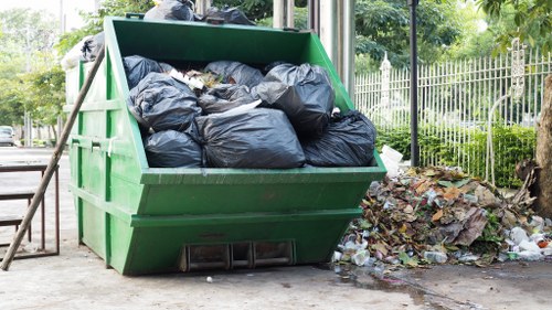 A large, green industrial waste skip filled with tightly packed black rubbish bags, situated outdoors on a paved surface near a residential area. The skip is partially lifted at the front, revealing the contents, and is positioned next to scattered litter and debris on the ground, with a background of green trees, fencing, and a lamppost, suggesting a typical municipal or private rubbish removal setting in a suburban environment. The scene is captured in daylight, emphasizing the textures of the plastic bags and the surrounding natural and urban elements. This image relates to rubbish removal services such as those provided by Harrow House Clearance aimed at clearing household or garden waste in London suburbs like Harrow.