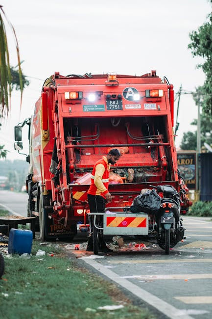 A green, wheeled rubbish skip filled with red bags of gardening or building materials, stacked inside a large container with a slightly rusted metal frame and a white section displaying contact inform